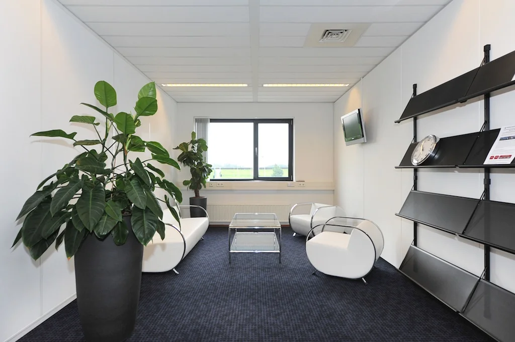 Moderne wachtruimte aan de Laan van 's-Gravenmade met witte designstoelen, grote planten en een wandrek met klok.